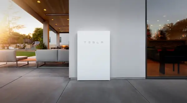 Un elegante Tesla Powerwall blanco se alza junto a una casa moderna con un patio y muebles de exterior.