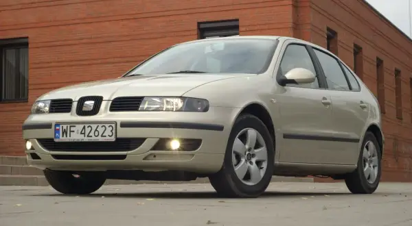 Jasny Seat Leon 1 z włączonymi światłami mijania i przeciwmgielnymi, zaparkowany przed ceglanym budynkiem.