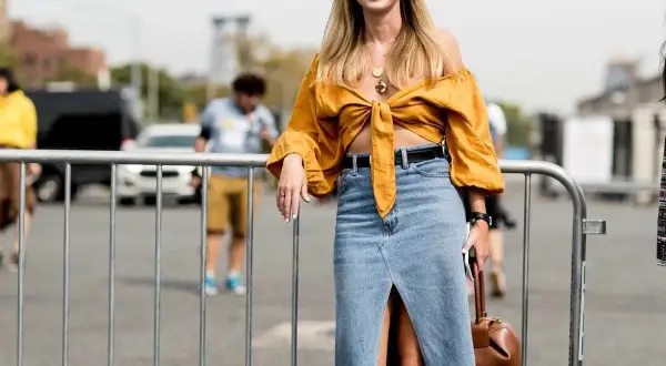 Mujer sonriente con un outfit falda vaquera larga, blusa amarilla anudada y bolso marrón.