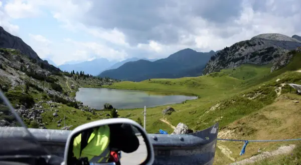 Widok z motocykla na malownicze **przełęcze alpejskie**, jezioro i góry pod zachmurzonym niebem.