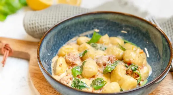 En skål med krämig gnocchi salsiccia, toppad med färsk basilika och parmesan. Citroner och en grå handduk i bakgrunden.