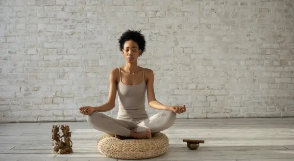 Mujer meditando en postura de loto, con los ojos cerrados y las manos en mudra. A su lado, una figura de bronce y un cuenco tibetano, elementos que invitan a la introspección y a la repetición de **palabras mantra**.