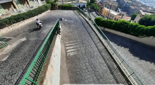 Le 13 discese di Napoli, una vista panoramica di una strada in discesa con un motorino, tra palazzi e vegetazione, che porta verso il mare.