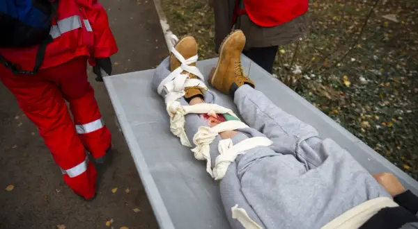 Na noszach leży osoba z opatrzonym złamaniem otwartym nogi. Ratownicy w czerwonych strojach udzielają pomocy.