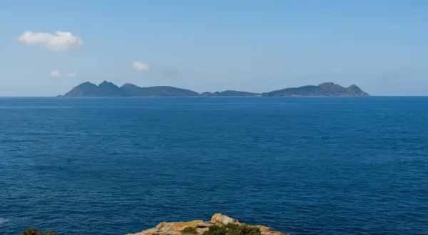 El mar azul se extiende hasta las islas de **cabo Estai**, con una costa rocosa y vegetación en primer plano.