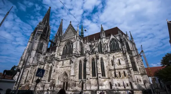 Der imposante Dom Regensburg ragt mit seinen Türmen in den blauen Himmel. Menschen und Autos bewegen sich auf dem Platz davor.