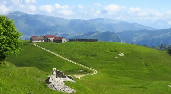 Un sentiero delle malghe serpeggia tra verdi colline, conducendo a un gruppo di rustici edifici con tetti rossi. In primo piano, ruderi di pietra e un piccolo stagno.