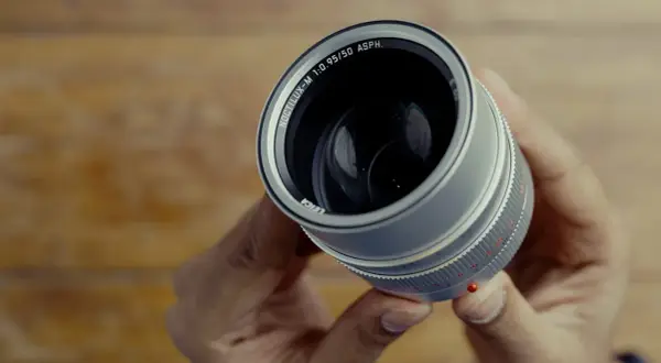 Hands hold a Leica Noctilux M 50 mm lens, its silver barrel and prominent f/0.95 aperture gleaming.