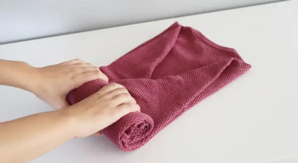 Dłonie zwijają różowy sweter, pokazując, jak składać swetry, by zajmowały mniej miejsca.