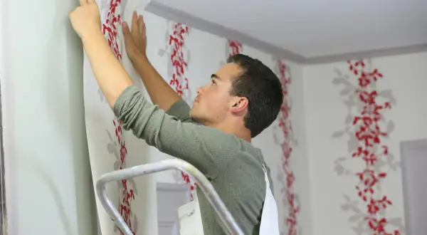 Mann steht auf Leiter und klebt Vinyltapete mit rotem Blumenmuster an die Wand.