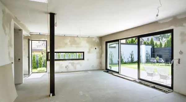 Wnętrze w trakcie budowy, ściany pokryte tynkiem gipsowo-wapiennym, gotowe do malowania. Duże okna wychodzą na ogród.