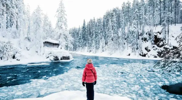 Une personne en manteau rouge admire un paysage hivernal de la pays Lapone, avec une rivière gelée et une cabane enneigée.