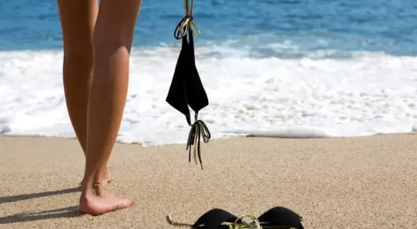 Nogi na piaszczystej plaży, obok czarny biustonosz. W tle błękitne morze i biała piana fal. Idealne miejsce na plażę nudystów.