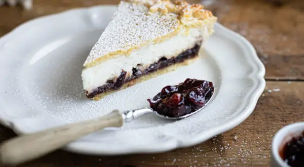Una fetta di crostata ricotta e visciole, cosparsa di zucchero a velo, su un piatto bianco. Un cucchiaino è pieno di visciole sciroppate.