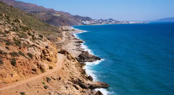 Costa rocosa con camino de tierra y el mar azul. Al fondo, se divisa la silueta de Mojácar y sus playas.