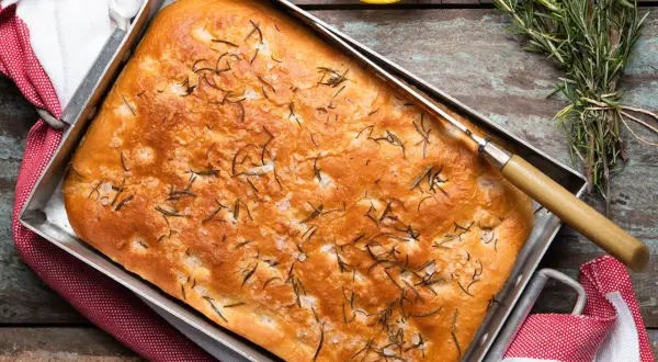En gyllene focaccia rosmarin i en plåt, toppad med örter och flingsalt. En kniv ligger på brödet, redo att skäras.