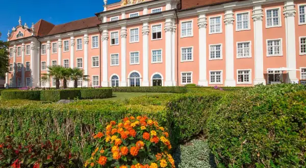 Das Meersburg Neues Schloss erstrahlt in Rosa vor blauem Himmel. Ein gepflegter Garten mit bunten Blumen und Palmen lädt zum Verweilen ein.