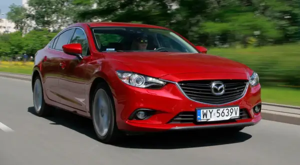 Czerwona Mazda 6 w ruchu ulicznym. Kierowca w okularach przeciwsłonecznych. Wiele pozytywnych mazda 6 opinie potwierdza jej sportowy charakter.