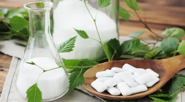 Ksylitol co to? Biały proszek w kolbie i słoiku, obok drewnianej łyżki z pastylkami. Liście brzozy sugerują naturalne pochodzenie.