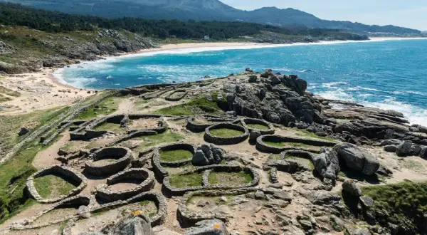 Ruinas circulares de piedra, vestigios de antiguos poblados, se extienden sobre un promontorio rocoso con el mar azul al fondo. Estos son algunos de los monumentos de Galicia.