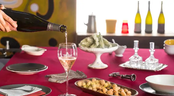 Versando vino tipico della Franciacorta in un calice, su un tavolo apparecchiato con stuzzichini e bottiglie.