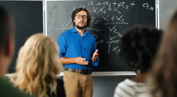 Nauczyciel w niebieskiej koszuli tłumaczy matematykę przy tablicy. Czy nauczyciel jest funkcjonariuszem publicznym? To pytanie nurtuje wielu.