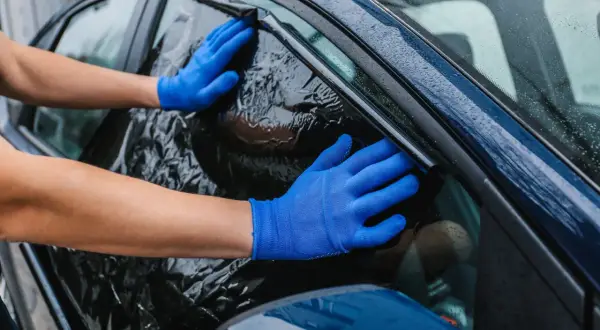 Ręce w niebieskich rękawiczkach usuwają pogniecioną folię z ciemnego okna samochodu. Poradnik: jak usunąć folię z okna.