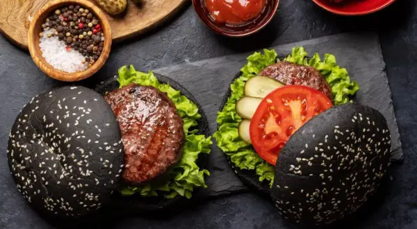 Dwa czarne burgery z sezamem, sałatą, pomidorem i ogórkiem kiszonym.