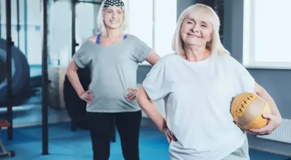 Dwie starsze kobiety ćwiczą na siłowni. Jedna trzyma piłkę. Może to być związane z tabletkami na menopauzę po których się chudnie.