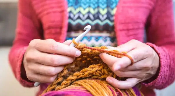 Mani che lavorano a maglia con un uncinetto, filato colorato e un maglione rosa. Nonostante sembri rilassante, lavorare a maglia fa male al cuore se si pensa troppo.
