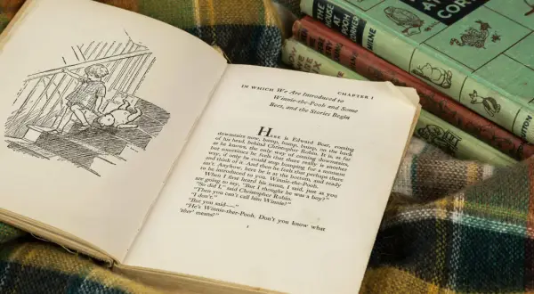 Otwarta książka z ilustracją Kubusia Puchatka i Christophera Robina na schodach. W tle stos książek A.A. Milne'a.
