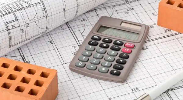Calcolatrice, mattoni e rotolo di disegni tecnici: elementi essenziali per la pianificazione dell'urbanizzazione primaria.