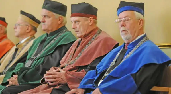 Czterech mężczyzn w togach akademickich i czapkach rektora siedzi w rzędzie.