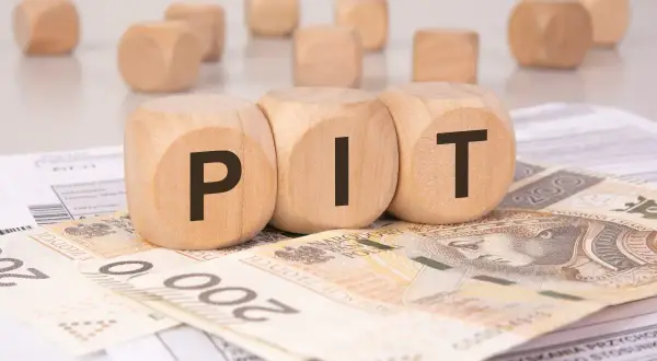 Drewniane klocki z literami "PIT" leżą na banknotach 200 zł i dokumentach podatkowych. To symboliczne przedstawienie podatku od darowizny.