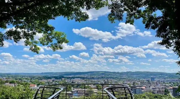 Blick vom Aussichtspunkt Wuppertal über die Stadt. Ein geschwungenes Geländer rahmt die weite Sicht auf Häuser und grüne Hügel unter einem blauen Himmel mit weißen Wolken ein.