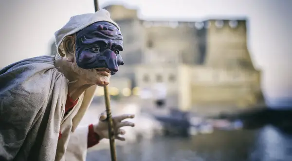 Un burattino di Pulcinella, con la sua maschera iconica, si staglia contro un castello sfocato. Una pulcinella storia che evoca tradizioni napoletane.