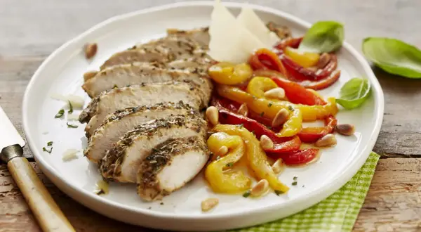 Sabroso pollo con pimientos asados, piñones y queso parmesano, servido en un plato blanco sobre una mesa de madera.