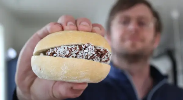 En hand håller upp en bulle med fyllning av choklad och kokosflingor. En man syns i bakgrunden. En riktig bulle med bulle!