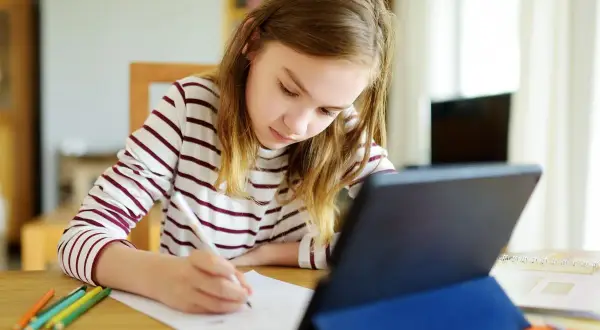 Dziewczynka w bluzie w paski pisze coś na kartce, obok tablet i kredki.