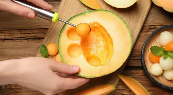 Ręka formuje kulki z melona, który ma niski melon indeks glikemiczny. Obok leżą kawałki melona i miseczka z owocowymi kulkami.