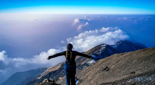 Eine Frau steht auf einem Berggipfel, die Arme ausgebreitet, und blickt auf eine Wolkendecke. Die Frage "wie hoch ist der Kilimandscharo?" hallt in der Weite wider.