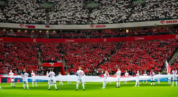 Piłkarze z flagą Polski na murawie, kibice tworzą biało-czerwone morze. Polska w rankingu FIFA zajmuje wysokie miejsce, co widać po dumie na trybunach.