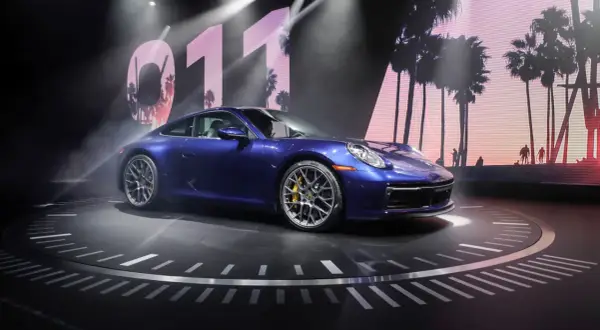 Un Porsche 911 992 azul metálico, con pinzas de freno amarillas, se exhibe sobre un escenario circular con un fondo de palmeras y el número 011.