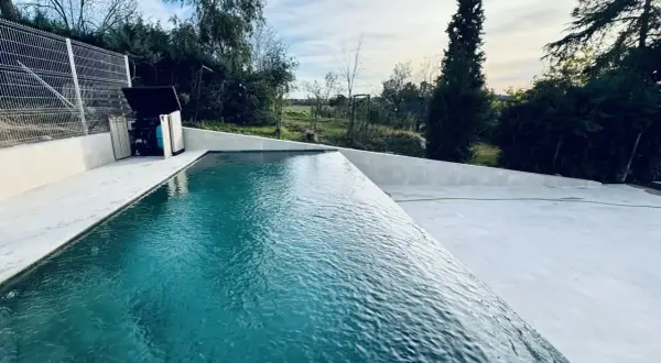 Una moderna piscina infinita con agua turquesa, rodeada de un área de concreto blanco y vegetación exuberante. El cielo azul con nubes blancas completa la escena.