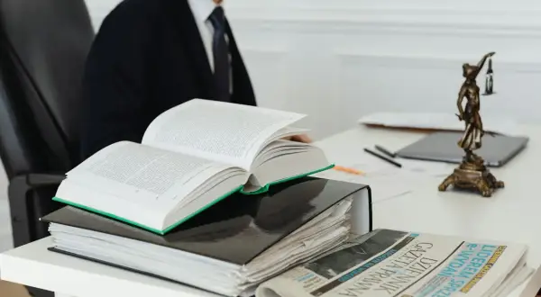 Otwarta księga prawnicza, akta i gazeta na biurku, obok statuetka Temidy.