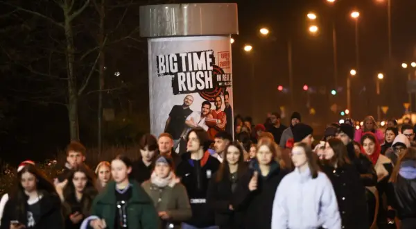 Tłum młodych ludzi zmierza na koncert Big Time Rush w Polsce. Plakat zespołu zdobi słup ogłoszeniowy.