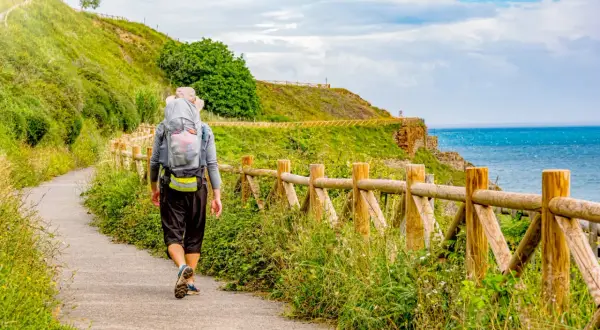 Vandrare på el camino de Santiago längs en kuststig med havet till höger och en grön kulle till vänster.