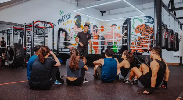 A boxing coach instructs a group sitting on the floor, preparing them for boxing reflex training. The gym features a boxing ring and punching bags.