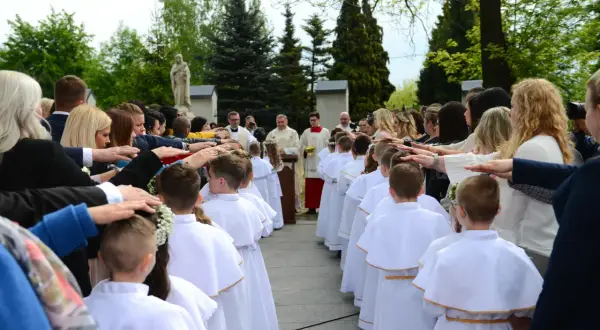 Dzieci w białych strojach komunijnych stoją w rzędzie, a dorośli kładą im ręce na głowach.