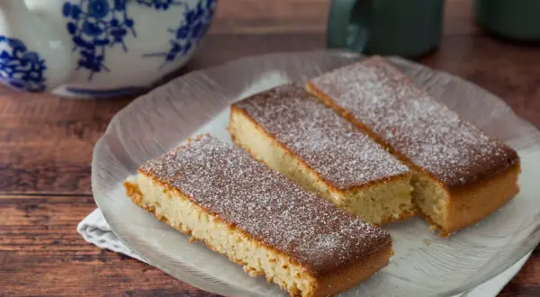 Saftiger mandelkuchen vom blech, bestäubt mit Puderzucker, auf einem Glas-Teller.